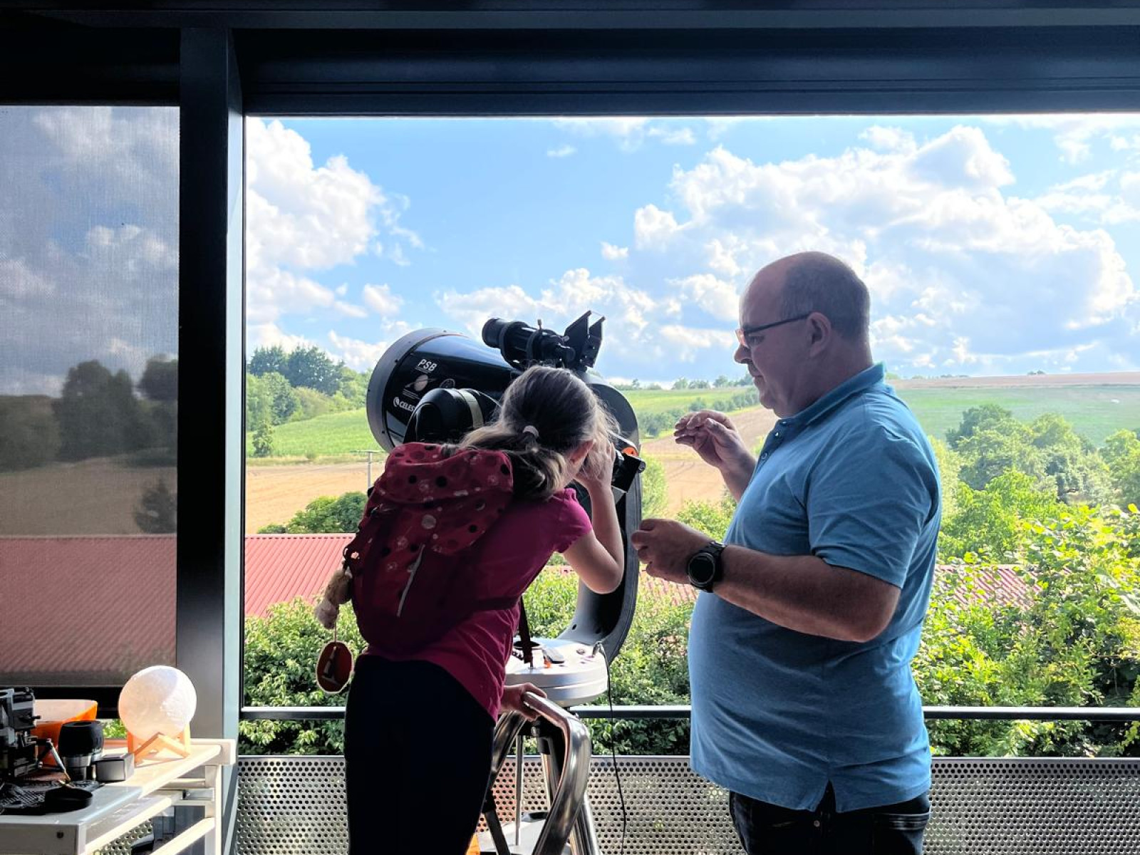 Ein Mann und ein Mädchen schauen durch ein Teleskop auf einem Balkon mit ländlichem Hintergrund.