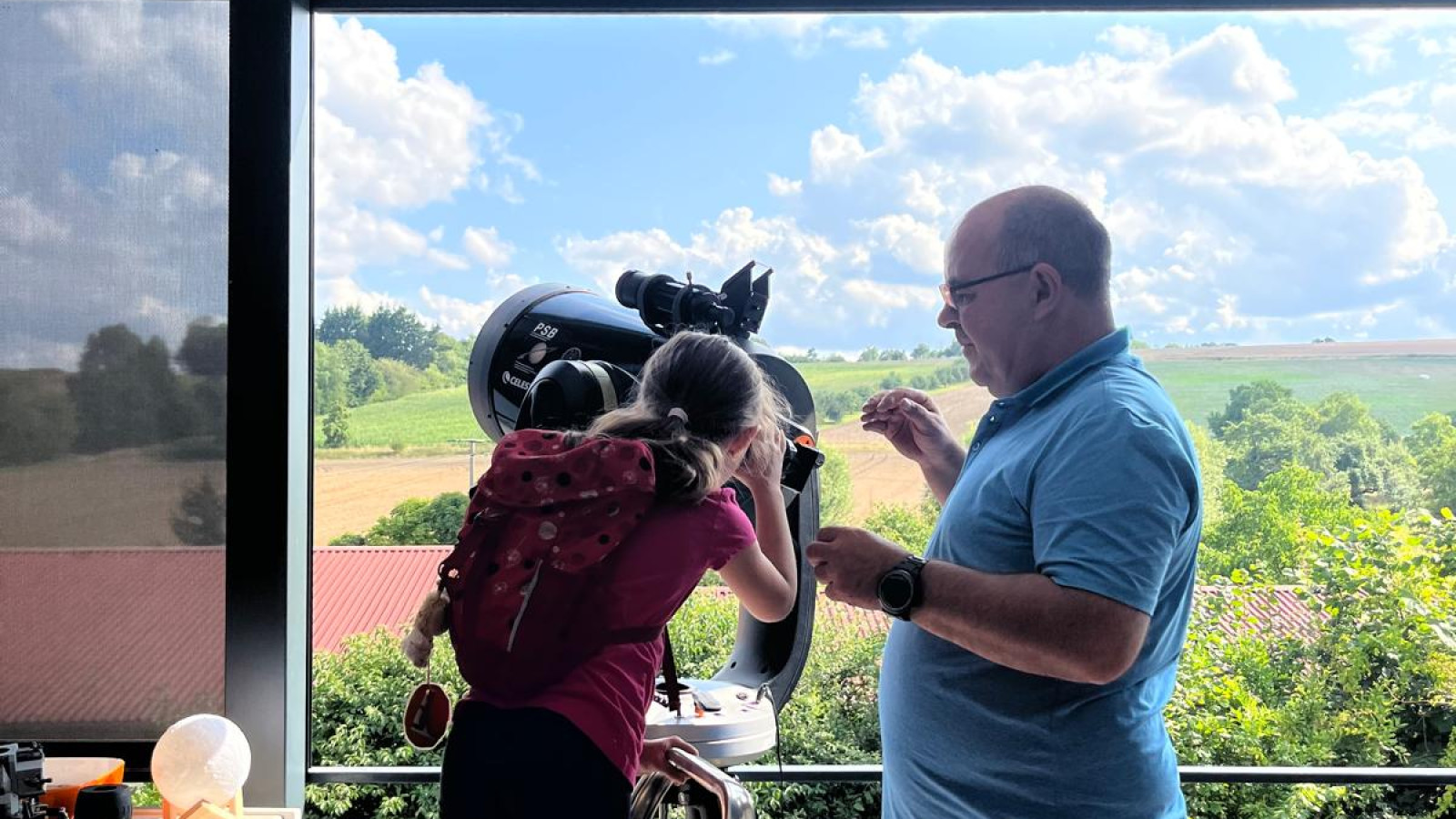 Mann und Kind auf einem Balkon mit Teleskop, Leiter und weiter Landschaft im Hintergrund.