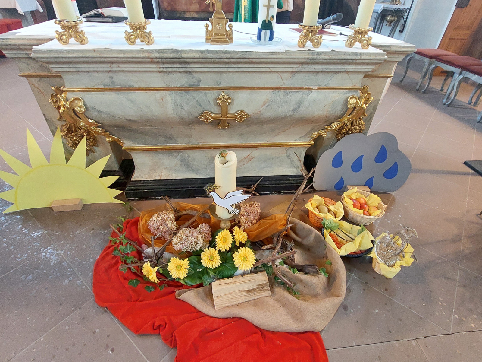 Altar mit Erntedankgaben, Sonnenblumen, Brot, Obst und Wetter-Symbolen in einer Kirche.
