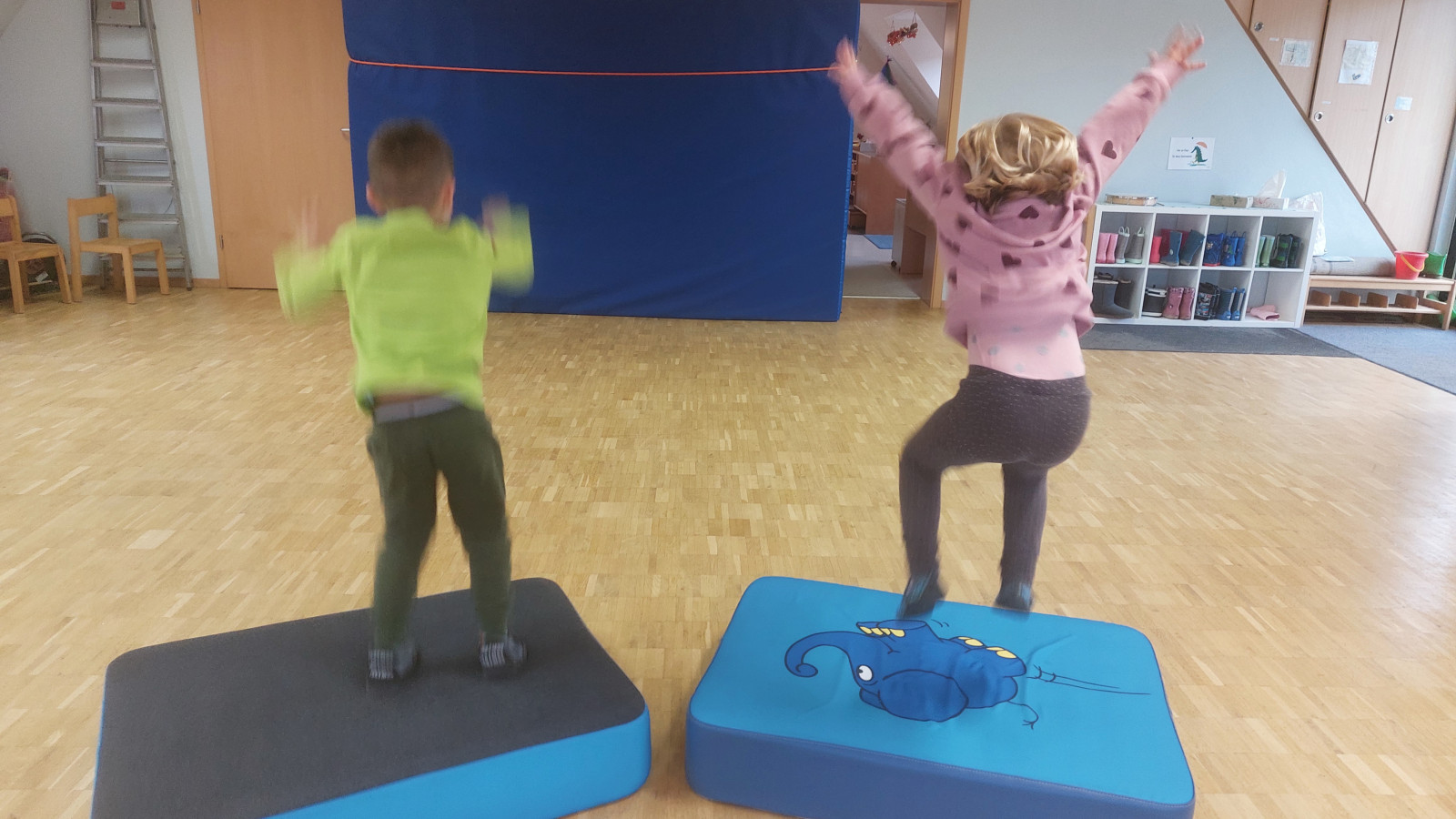 Zwei Kinder springen fröhlich auf blauen und grauen Matten in einem Raum mit Holzboden.