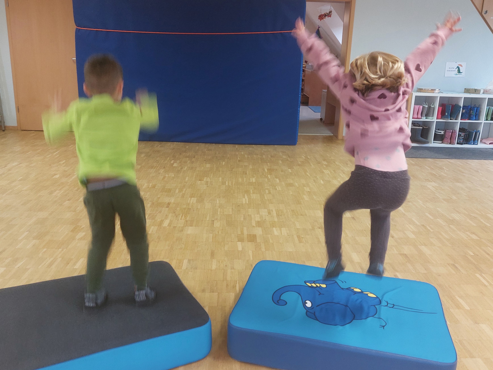 Zwei Kinder springen von blauen Matten in einer Turnhalle mit Holzboden.