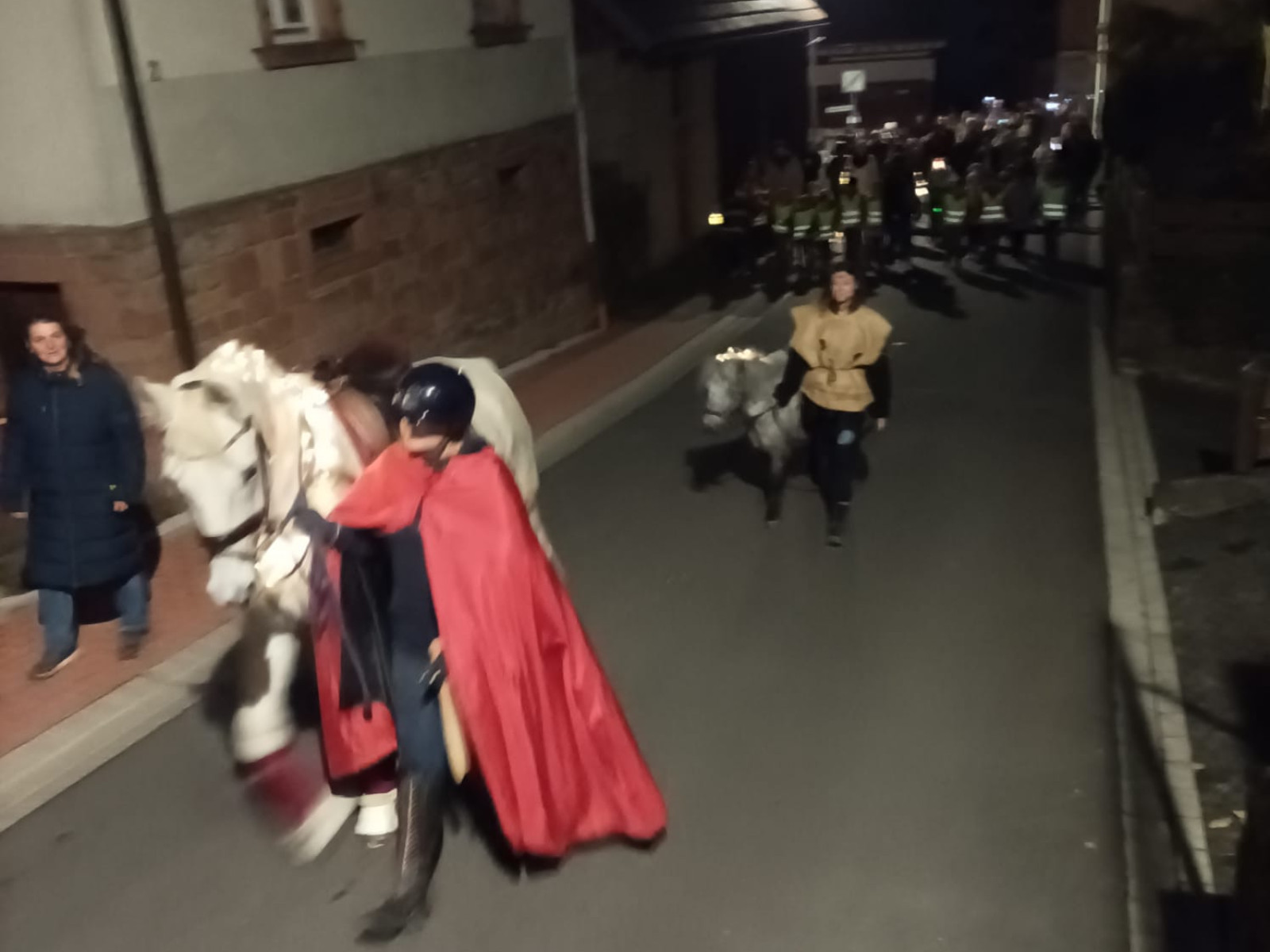 Nachtszene eines Reiters im roten Umhang auf einem weißen Pferd, gefolgt von einem Laternenumzug.