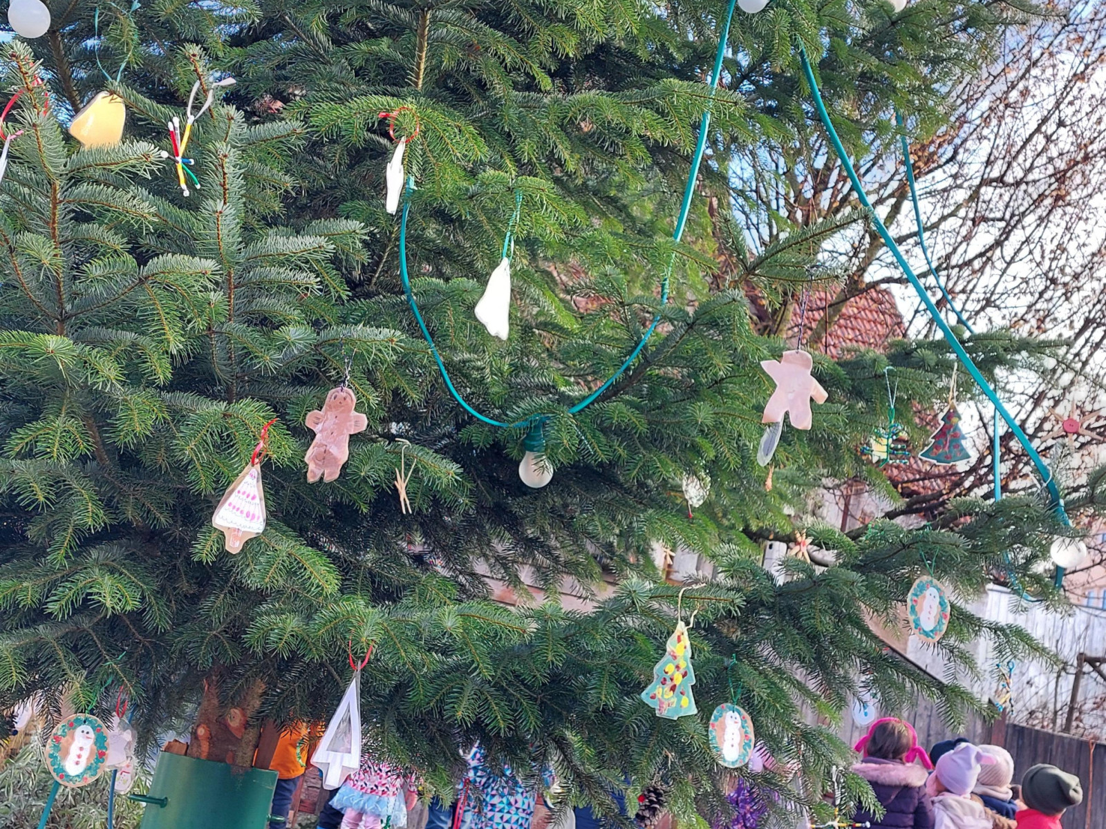 Kinder stehen um einen großen Weihnachtsbaum herum und hängen Dekorationen im Freien auf.