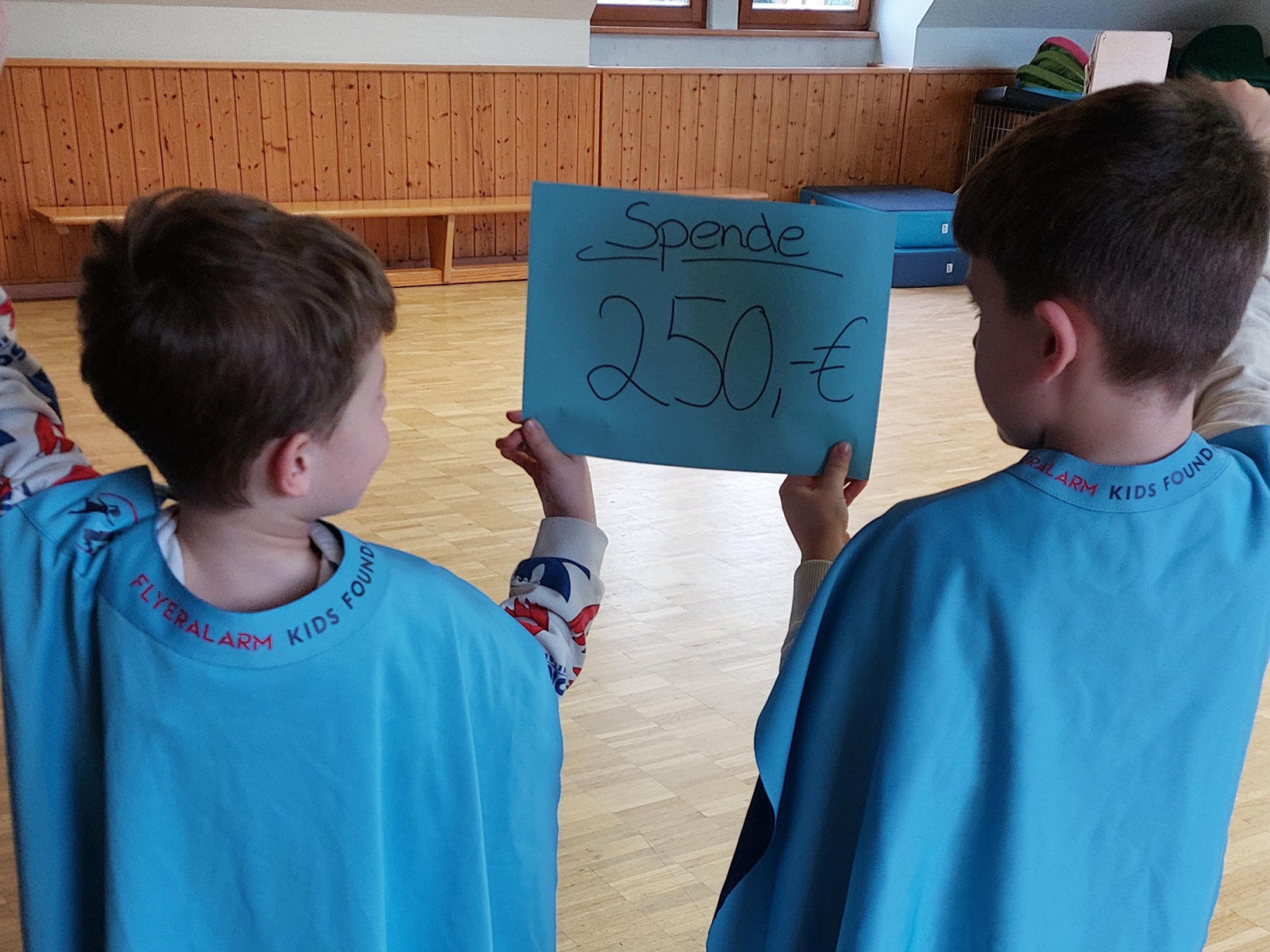 Zwei Kinder in blauen Capes halten ein Spendenplakat mit '250€' in einem hellen Raum.