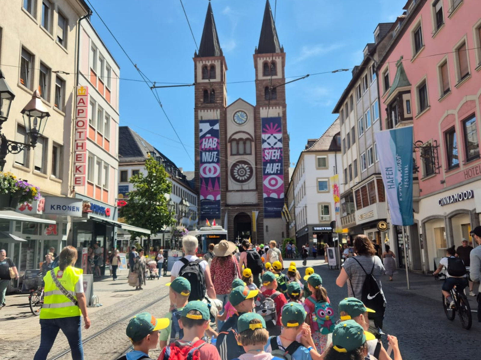 Kindergruppe in farbigen Hüten und Rucksäcken geht bei Sonnenschein durch eine belebte Straße, mit einer Kirche im Hintergrund.