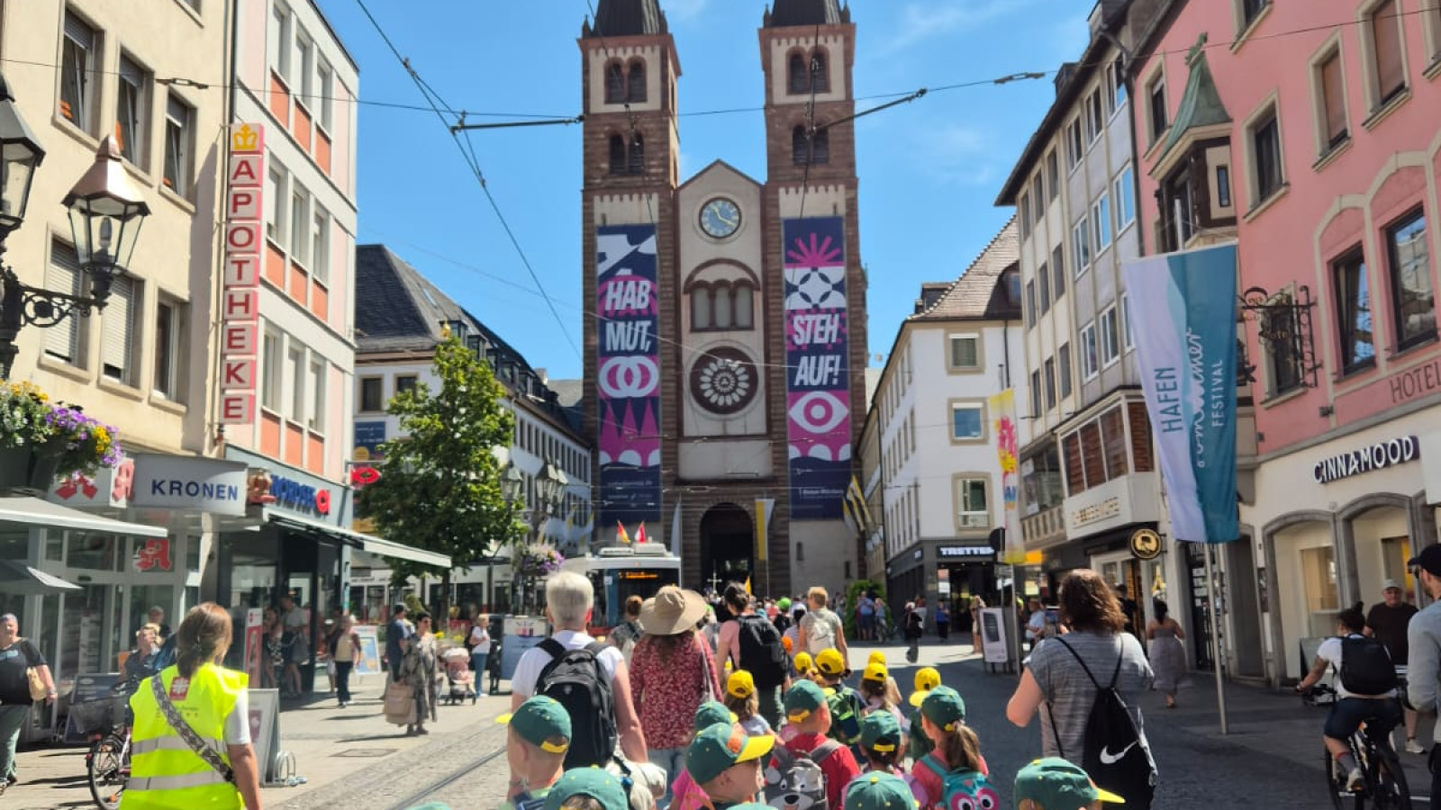 Kindergruppe mit grünen Kappen vor einer Kirche mit zwei Türmen in einer Stadtstraße.