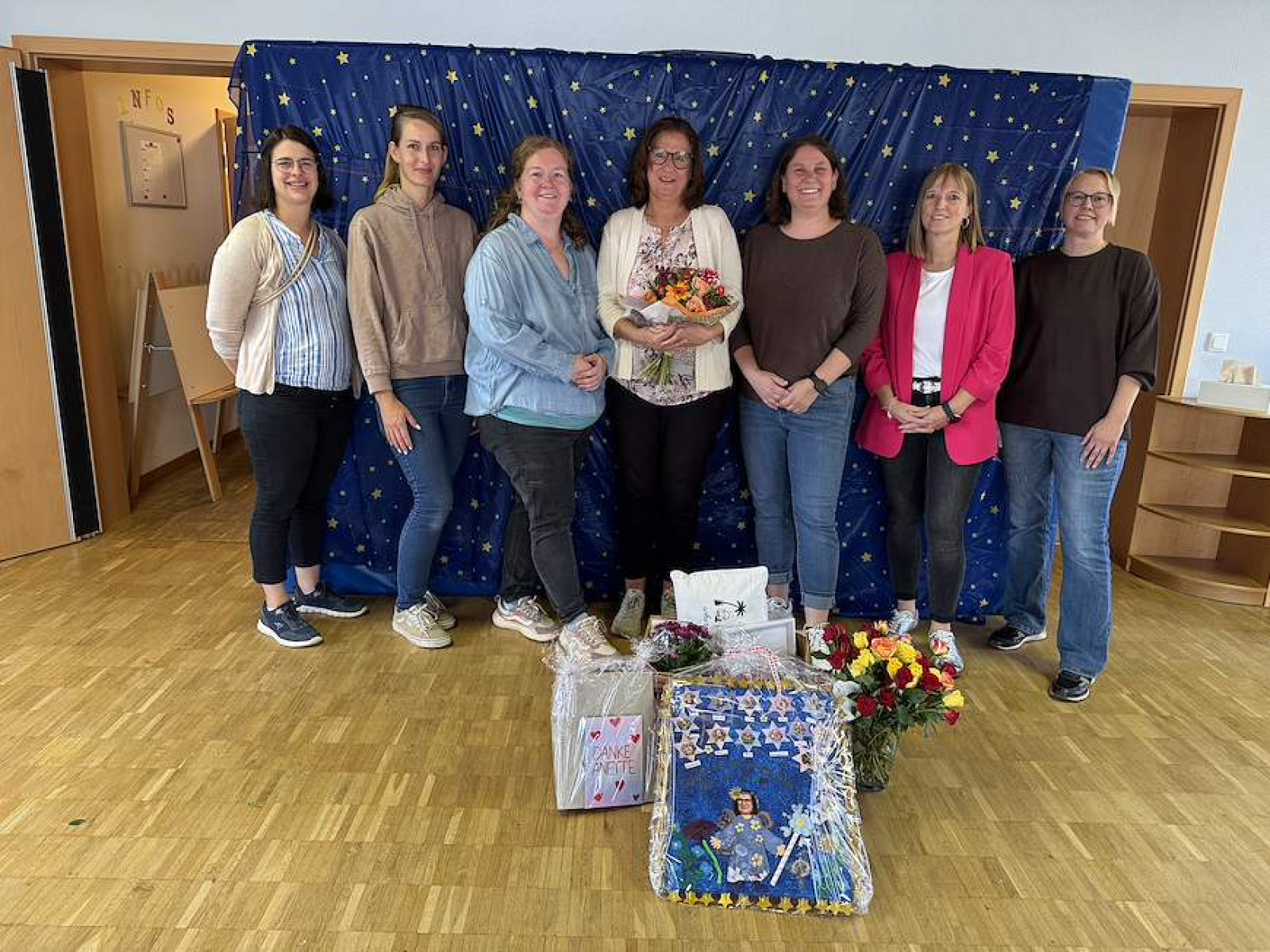 Sieben Personen vor einem Sternenhintergrund mit Blumen und Geschenken auf dem Boden.
