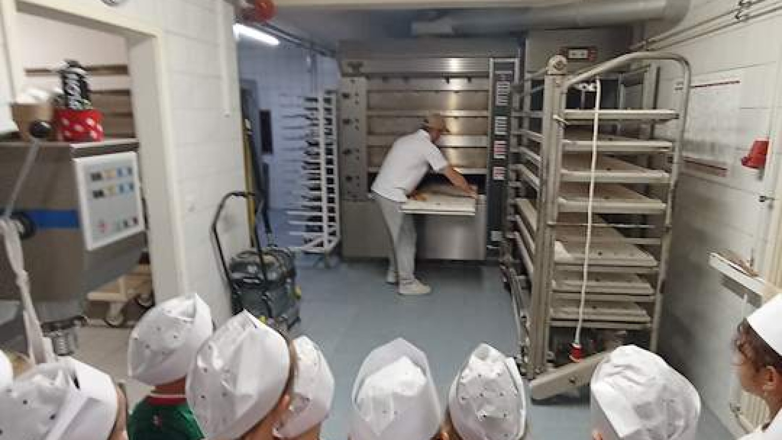 Kinder mit Kochmützen beobachten einen Bäcker in einer Bäckerei.