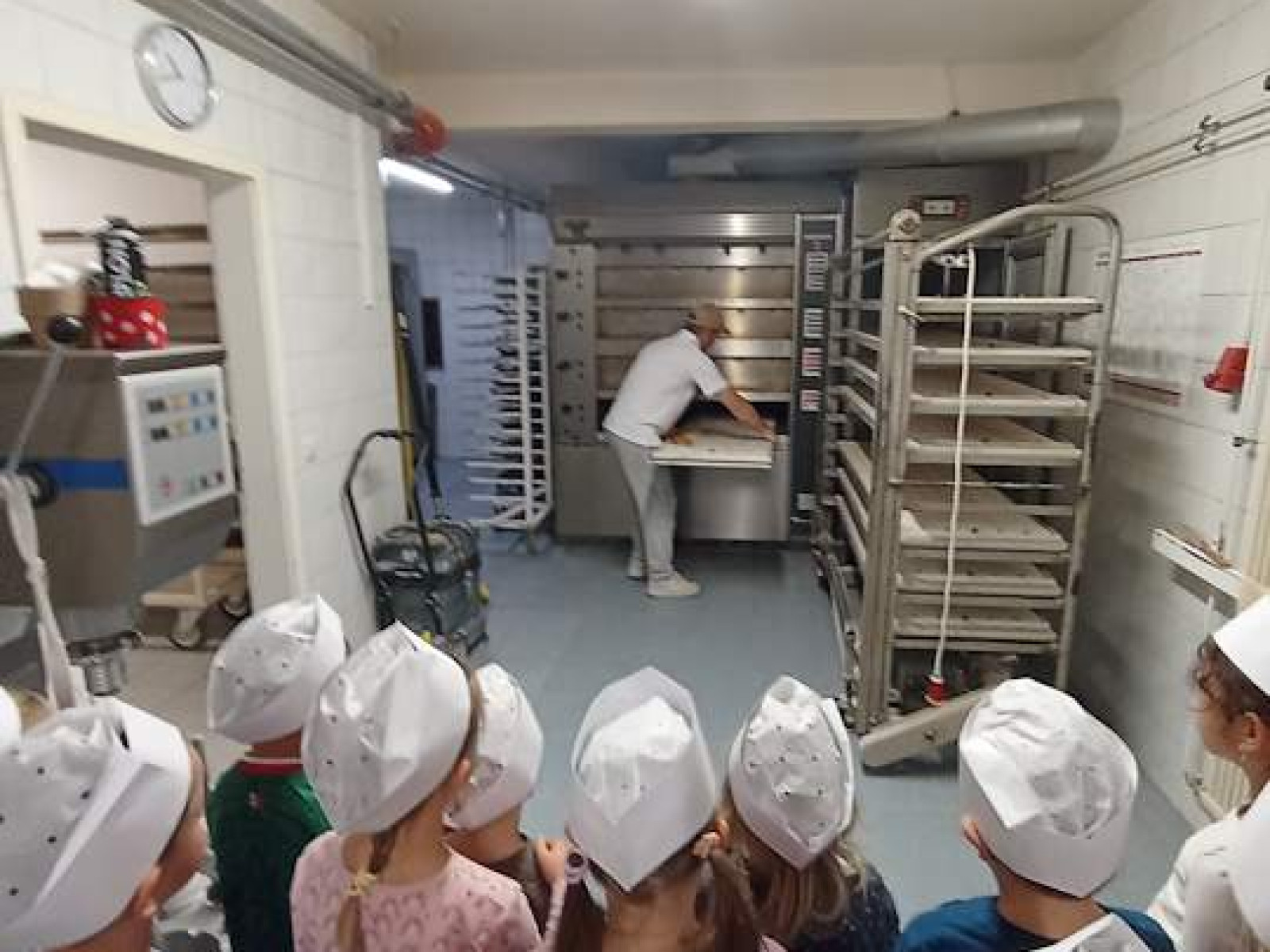 Kinder mit weißen Mützen schauen einem Bäcker in einer Bäckerei zu.